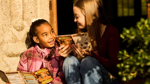 Two girls sitting together in a sunny doorway at Lanhydrock, smiling and exploring the illustrated Weave a Wild Tale autumn activity leaflets.